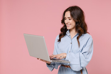 Smiling beautiful young brunette woman 20s wearing casual blue shirt dress posing standing hold in hands working in laptop pc computer isolated on pastel pink colour background, studio portrait. - obrazy, fototapety, plakaty