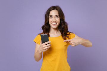 Cheerful young brunette woman 20s wearing basic yellow t-shirt pointing index finger on mobile cell phone typing sms message looking camera isolated on pastel violet colour background studio portrait.