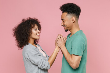 Side view of smiling young african american couple two friends guy girl in gray green casual clothes posing holding hands looking at each other isolated on pastel pink wall background studio portrait.