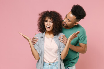 Angry irritated displeased young african american couple two friends guy girl in gray green clothes posing hugging spreading hands swearing isolated on pastel pink color background studio portrait.