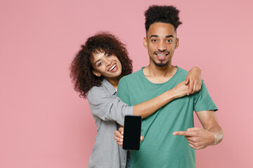 Funny african american couple friends guy girl in gray green clothes point index finger on mobile phone with blank empty screen mock up copy space isolated on pastel pink background studio portrait.