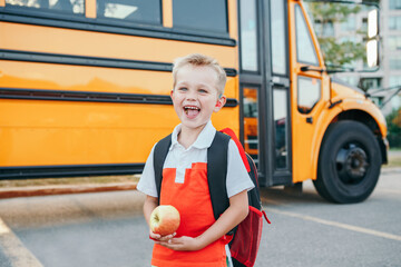 Caucasian boy student with backpack near yellow bus on first September day. Child kid eating apple...