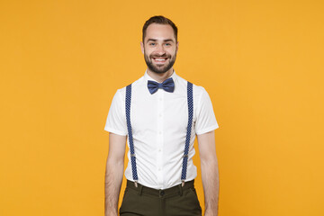 Smiling handsome cheerful funny young bearded man 20s wearing white shirt bow-tie suspender posing standing and looking camera isolated on bright yellow color wall background, studio portrait.