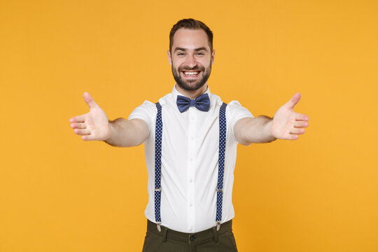 Smiling pleasant joyful young bearded man 20s in white shirt bow-tie suspender posing standing reach out stretch hands looking camera isolated on bright yellow color wall background studio portrait.