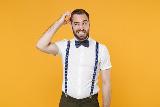 Preoccupied concerned puzzled young bearded man 20s in white shirt bow-tie suspender posing standing put hand on head looking aside isolated on bright yellow color wall background studio portrait.