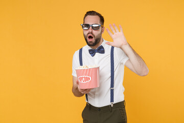 Shocked scared young bearded man 20s wearing white shirt bow-tie suspender 3d glasses watching movie film, holding bucket of popcorn covering with hand isolated on yellow background studio portrait.