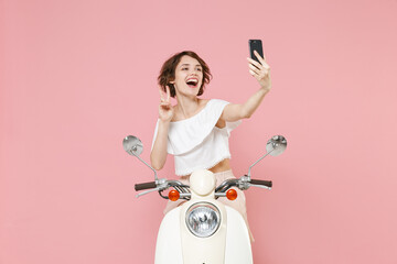Funny young brunette woman 20s wearing white summer clothes doing selfie shot on mobile phone showing victory sign sitting and driving moped isolated on pastel pink colour background, studio portrait.