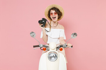 Excited young brunette woman 20s wearing white summer clothes hat eyeglasses hold in hand retro vintage photo camera sitting driving moped isolated on pastel pink colour background studio portrait.