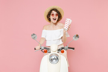 Excited young brunette woman 20s wearing white summer clothes hat eyeglasses hold in hand plastic cup of cola or soda sitting driving moped isolated on pastel pink colour background studio portrait.