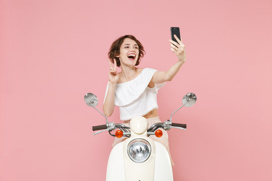 Funny young brunette woman 20s wearing white summer clothes doing selfie shot on mobile phone showing victory sign sitting and driving moped isolated on pastel pink colour background, studio portrait.