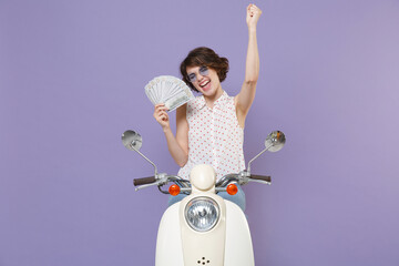 Happy young brunette woman 20s in white dotted shirt glasses hold fan of cash money in dollar banknotes doing winner gesture driving moped isolated on pastel violet colour background studio portrait.