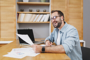 Cheerful smiling funny young bearded business man 20s wearing blue shirt glasses sitting at desk with papers document working on laptop pc computer writing in notebook at home or office.