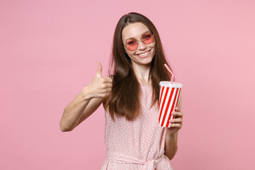 Smiling young brunette woman 20s wearing pink summer dotted dress eyeglasses posing hold plastic cup of cola or soda showing thumb up isolated on pastel pink color wall background studio portrait.