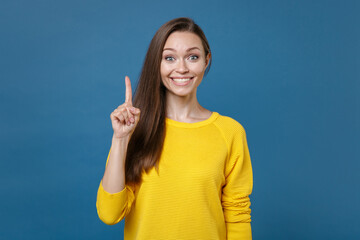 Smiling excited young brunette woman 20s wearing yellow casual clothes posing standing holding index finger up with great new idea looking camera isolated on blue color background, studio portrait.