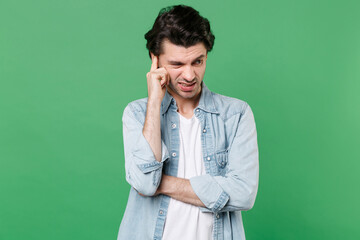 Fototapeta na wymiar Preoccupied puzzled confused young brunet man 20s wearing casual clothes white t-shirt denim shirt posing standing put hand on head looking aside isolated on green color background studio portrait.