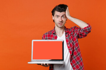 Perplexed puzzled young man 20s in white t-shirt red checkered shirt hold laptop pc computer with blank empty screen mock up copy space put hand on head isolated on orange background studio portrait.