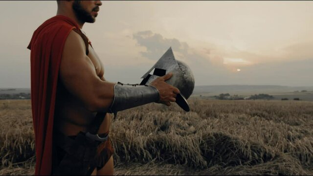 Shirtless spartan wearing helmet in dry field, sunset.