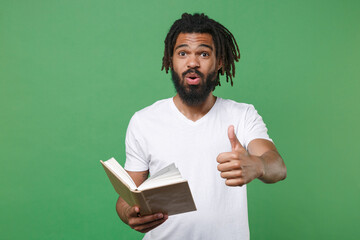 Shocked amazed young african american man guy with dreadlocks 20s wearing white casual t-shirt posing holding in hand reading book showing thumb up isolated on green color background studio portrait.