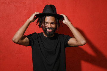Smiling handsome attractive young african american man guy with dreadlocks 20s in black casual t-shirt posing hold hat on head looking camera isolated on bright red color background studio portrait.