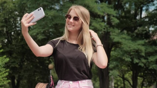Beautiful blonde girl with a smartphone makes a selfie. Summer outdoors girl blogger posing in front of smartphone cameras.