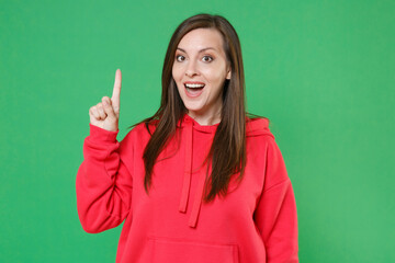 Excited young brunette woman 20s wearing bright red casual streetwear hoodie posing holding index finger up with great new idea looking camera isolated on green color wall background studio portrait.