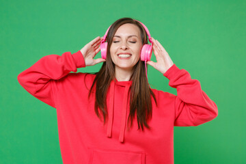 Smiling young brunette woman 20s in bright red casual streetwear hoodie posing listening music with headphones put hands on head keeping eyes closed isolated on green color background studio portrait.