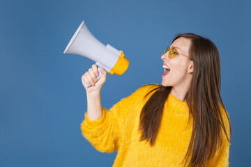 Excited beautiful cheerful funny young brunette woman 20s wearing yellow fur sweater eyeglasses posing screaming in megaphone looking aside up isolated on blue color wall background studio portrait.