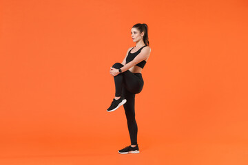 Fototapeta na wymiar Full length side view portrait young fitness sporty woman in black sportswear posing training working out doing stretching exercising for legs looking aside isolated on orange color background studio.