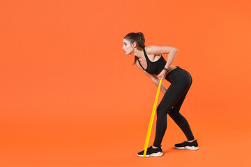 Full length side view portrait of young fitness sporty woman in black sportswear posing training working out doing exercises with fitness gums looking aside isolated on orange color background studio.