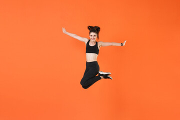 Fototapeta na wymiar Full length portrait of smiling funny young fitness sporty woman wearing black sportswear posing training working out jumping spreading hands looking camera isolated on orange color background studio.