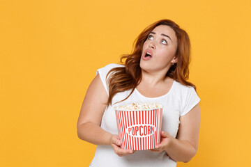 Shocked young redhead plus size body positive female woman girl 20s in white t-shirt watching movie film, hold bucket of popcorn looking aside up isolated on yellow color background studio portrait.