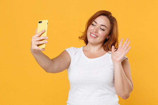 Smiling young redhead plus size body positive female woman girl in white casual t-shirt doing selfie shot on mobile phone waving greeting with hand isolated on yellow color background studio portrait.