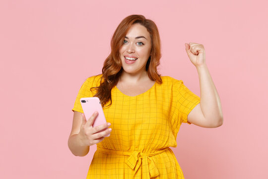 Happy young redhead plus size body positive female woman girl 20s in yellow dress posing using mobile cell phone doing winner gesture looking camera isolated on pastel pink background studio portrait.