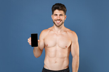 Smiling young fitness sporty guy bare-chested muscular sportsman isolated on blue background. Workout sport motivation lifestyle concept. Mock up copy space. Hold mobile phone with blank empty screen.