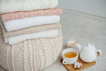 Fototapeta na wymiar A stack of knitted sweaters, a mug of hot tea, a teapot, a wooden stand on an ottoman. Cozy autumn. Winter breakfast. Roses. Candle.