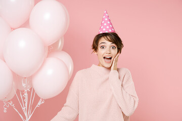 Excited young brunette woman in knitted casual sweater birthday hat isolated on pastel pink background. Birthday holiday party people emotions concept. Celebrating hold air balloons put hand on cheek.