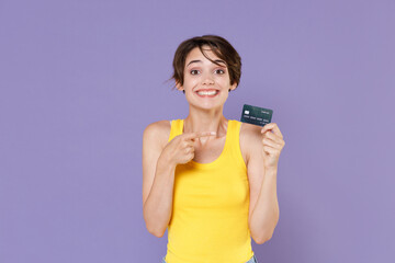 Smiling young brunette woman girl in yellow casual tank top posing isolated on pastel violet background studio portrait. People emotions lifestyle concept. Pointing index finger on credit bank card.