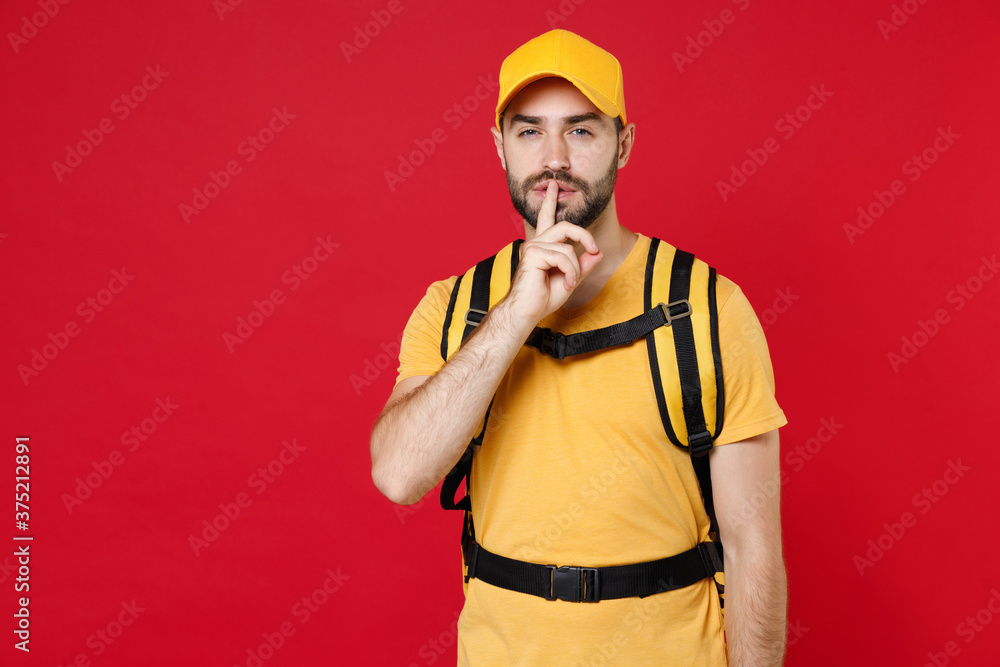 Wall mural Delivery employee man guy male 20s in yellow cap t-shirt uniform thermal bag backpack with food work as courier isolated on red background studio. Service during quarantine coronavirus covid-19 virus.