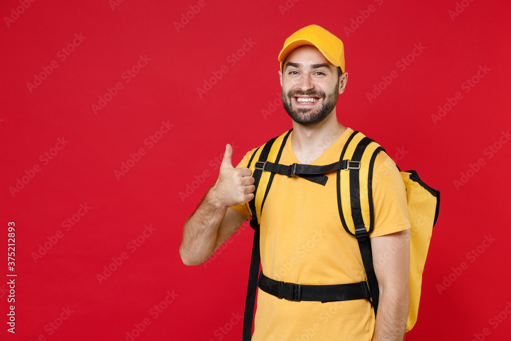 Wall mural Delivery employee man guy male 20s in yellow cap t-shirt uniform thermal bag backpack with food work as courier isolated on red background studio. Service during quarantine coronavirus covid-19 virus.