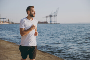 Portrait of handsome attractive young bearded athletic man guy 20s in casual white t-shirt black shorts posing training running looking aside at sunrise over the sea outdoors.