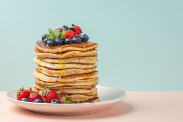 A stack of pancakes with honey, strawberries and blueberries. Copy space, text box. Light blue and pink background.