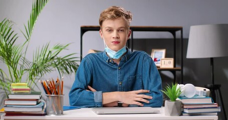 Portrait of Caucasian teenage boy sitting at desk with books and textbooks, taking off medical mask and smiling to camera. Teenager schoolboy. Homework concept. School life. Pandemic concept. - Powered by Adobe