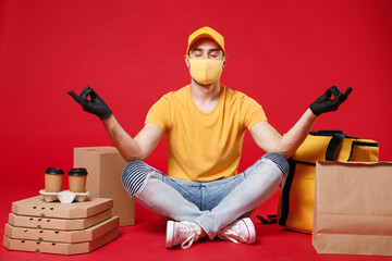 Delivery employee man in yellow cap face mask gloves tshirt sit at thermal takeaway food bag backpack pizza boxes work courier service during quarantine coronavirus covid-19 isolated on red background