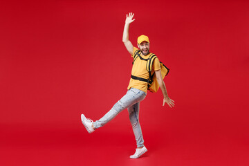 Full length delivery employee man guy male 20s in yellow cap t-shirt uniform thermal food bag backpack work courier service during quarantine covid-19 virus, standing isolated on red background studio