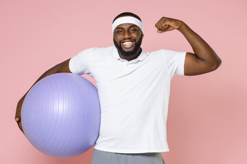 Smiling young bearded african american fitness sports man in white headband t-shirt hold fitball showing biceps muscles spending time in gym isolated on pastel pink color background studio portrait.
