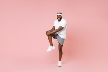 Full length portrait of smiling young african american fitness sports man 20s in headband t-shirt doing stretching exercising for legs spending time in gym isolated on pink color background studio. - Powered by Adobe