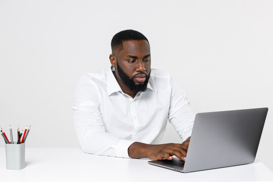 Serious handsome young african american male business man in classic white shirt posing working in office sitting at desk using laptop pc computer isolated on white color wall background.