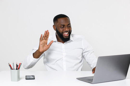 Smiling young african american male business man in white shirt posing work in office sit at desk using laptop pc computer making video call waving greeting with hand isolated on white background.