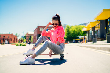 Cheerful female teenager in stylish sunglasses and streetwear resting at longboard during sport...