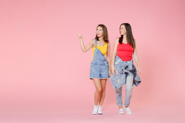Obraz premium Full length portrait of two smiling young brunette women friends 20s wearing casual denim clothes pointing index finger aside on mock up copy space isolated on pastel pink colour background studio.